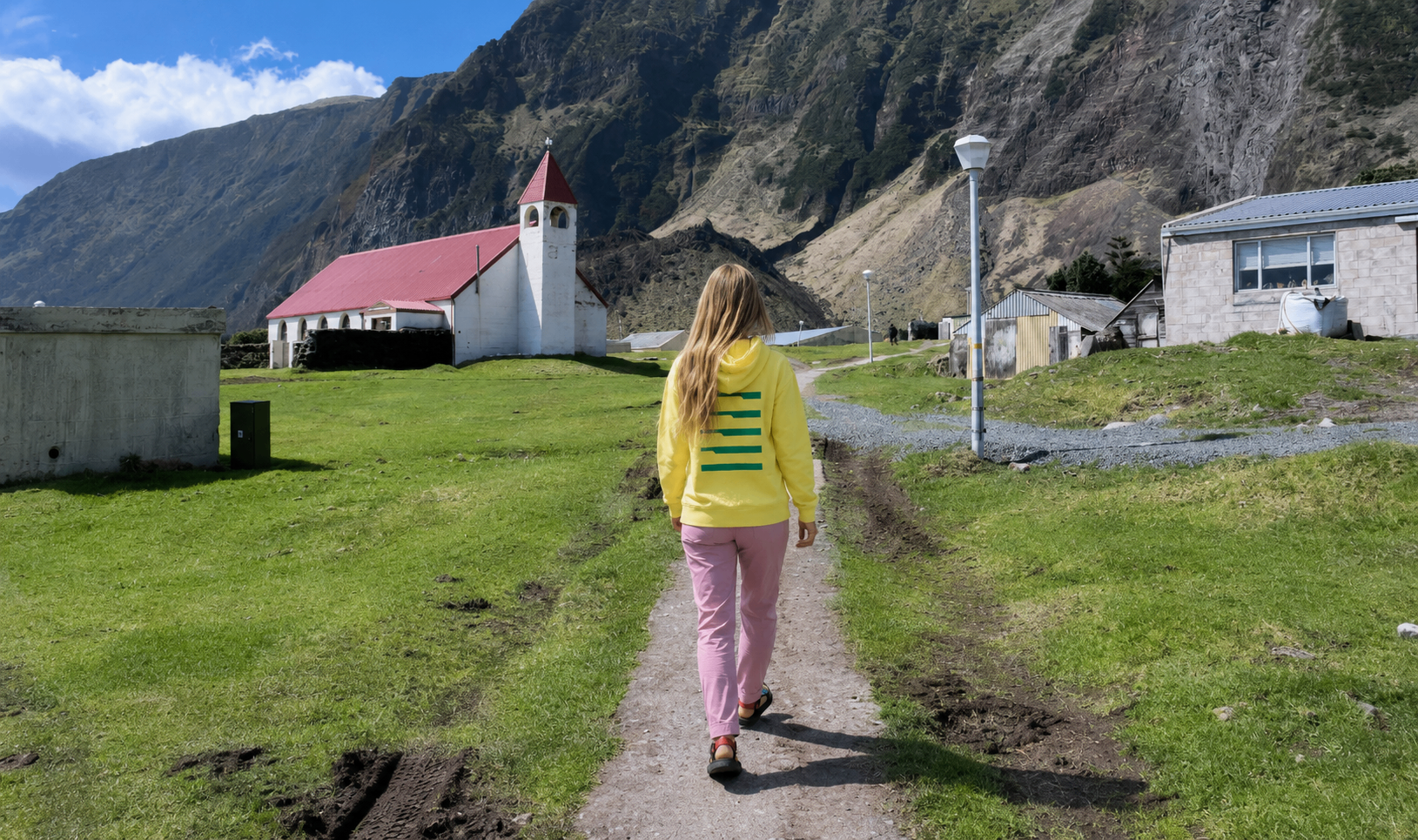A person in a bright yellow hoodie walking toward a small white church with a red roof, nestled beneath the towering volcanic cliffs of Tristan da Cunha