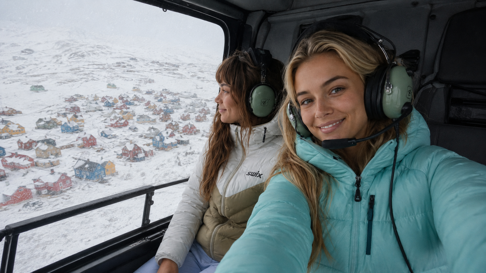 Vera and Mila in a helicopter cockpit looking down at snowy Greenland landscape with colorful houses below